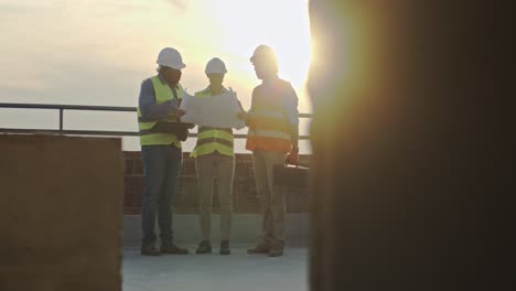 Caucasian-woman-boss-with-laptop-computer-standing-on-the-sunset-on-the-roof-with-two-male-multiethnic-builders.